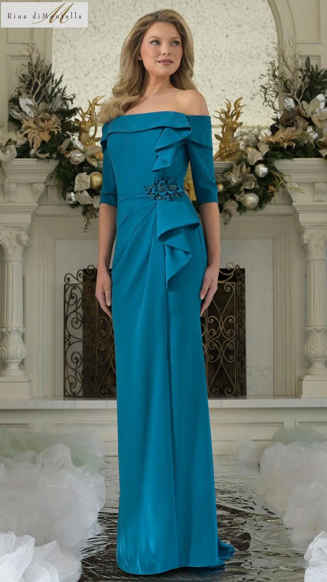 A woman stands before a decorated fireplace wearing the teal blue Rina Di Montella RD3202 long formal dress, featuring an off-the-shoulder design with ruffle and floral embellishments.